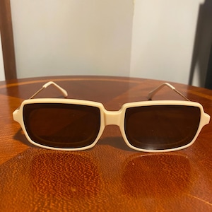 May include: Cream-colored sunglasses with a rectangular frame and brown lenses. The sunglasses have thin, gold-colored arms. The frame is a light beige color. The sunglasses are resting on a wooden surface.