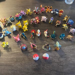 Può includere: Una collezione di figurine Pokémon colorate, tra cui Pikachu, Charmander e Bulbasaur, è disposta su una superficie di legno. Le figure variano per dimensioni e posa, mostrando una varietà di personaggi del popolare franchise. Sono presenti anche due Poké Ball rosse e bianche.