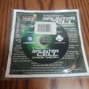 Op de afbeelding: Een verzegelde kopie van de videogame "Tom Clancy's Splinter Cell: Chaos Theory" voor Xbox. De schijf is zichtbaar door de transparante plastic verpakking. Het logo en de titel van het spel staan prominent op de schijf en de verpakking.