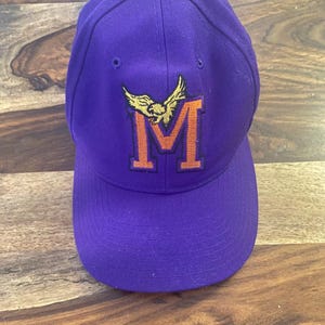 May include: A purple baseball cap with a curved peak. The cap has an embroidered design of a golden bird above an orange letter "M". The cap is made of a durable fabric and has a structured fit.
