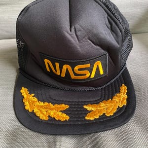 May include: A black trucker hat featuring a rectangular patch with the yellow embroidered text NASA on the front panel. The brim is decorated with two yellow embroidered oak leaf clusters, and the back panels are made of black mesh material.