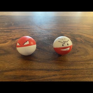 Può includere: Due giocattoli Poké Ball bianchi e rossi, uno con un design rosso e bianco e l'altro con una faccia arrabbiata disegnata. I giocattoli sono su una superficie di legno.