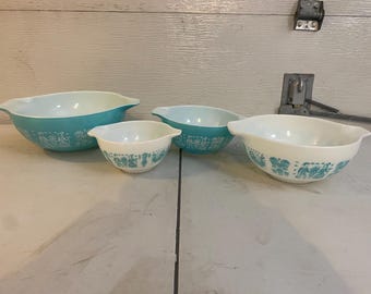May include: A set of four white and turquoise glass mixing bowls with integrated handles. The bowls feature a repeating pattern of figures in colonial-style clothing printed in turquoise around the exterior. The set includes one large bowl, two medium bowls, and one small bowl.