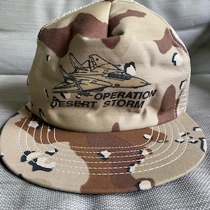 Peut inclure: Une casquette de type trucker à motif camouflage beige, marron et crème, ornée d'une broderie représentant un avion de chasse et du texte noir "OPERATION DESERT STORM" sur le panneau avant. Elle possède une visière plate et un filet arrière.