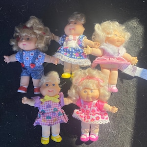 May include: Five vintage doll figures with blonde curly hair and rosy cheeks. The dolls wear colorful dresses and outfits, including denim overalls, floral prints, and a pink dress with a white collar. Each doll has small shoes in various colors.