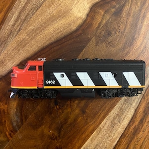 May include: A model train locomotive with a red front and black body. The train features white and black diagonal stripes along the side and the number 9162. The wheels and undercarriage are black and the train sits on a wooden surface.