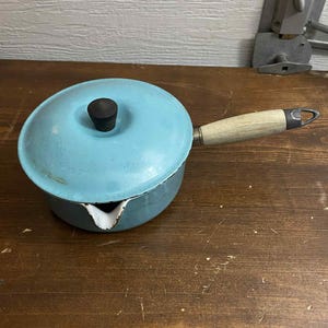 May include: A vintage, light blue enamel saucepan with a matching lid and a wooden handle. The saucepan has a spout for pouring and a black knob on the lid. The handle is light-colored wood. The pot is on a wooden surface.