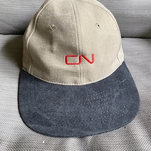 May include: A baseball cap featuring a tan crown and a dark grey brim. The front of the crown displays the letters CN embroidered in red thread. The cap includes a small dark grey button at the top centre and stitched eyelets on the panels.