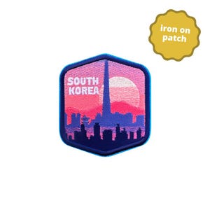 May include: An iron-on patch featuring a pink and purple sunset over a cityscape with a tall tower in the center. The text "SOUTH KOREA" is embroidered in white on the patch. The patch is shaped like a hexagon with a blue outline.