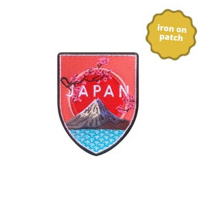 May include: Embroidered patch depicting a mountain scene with the word "JAPAN" in white. The patch has a red background with a mountain, water, and cherry blossoms. The patch is shield-shaped with a yellow "iron on patch" tag.