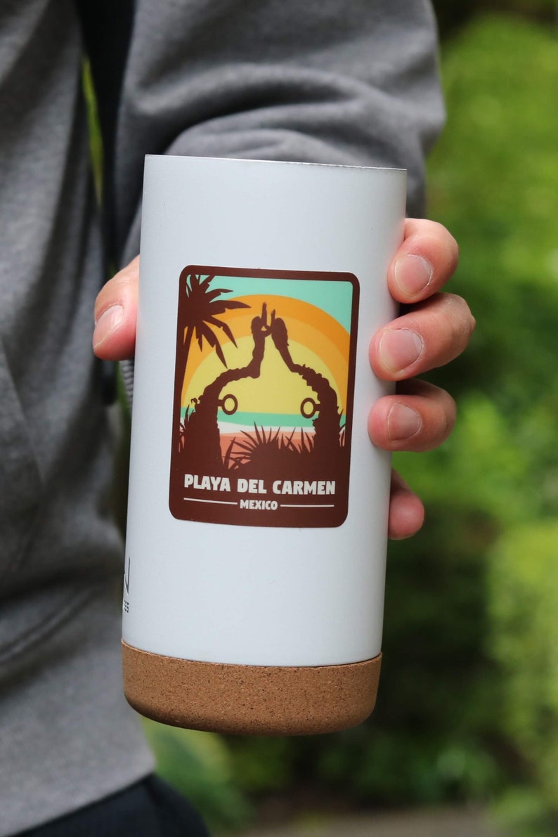 Pode incluir: Caneca de viagem branca com fundo de corti&ccedil;a e um adesivo castanho com a silhueta de duas pessoas em p&eacute; numa praia com uma palmeira e o texto "Playa del Carmen - M&eacute;xico".