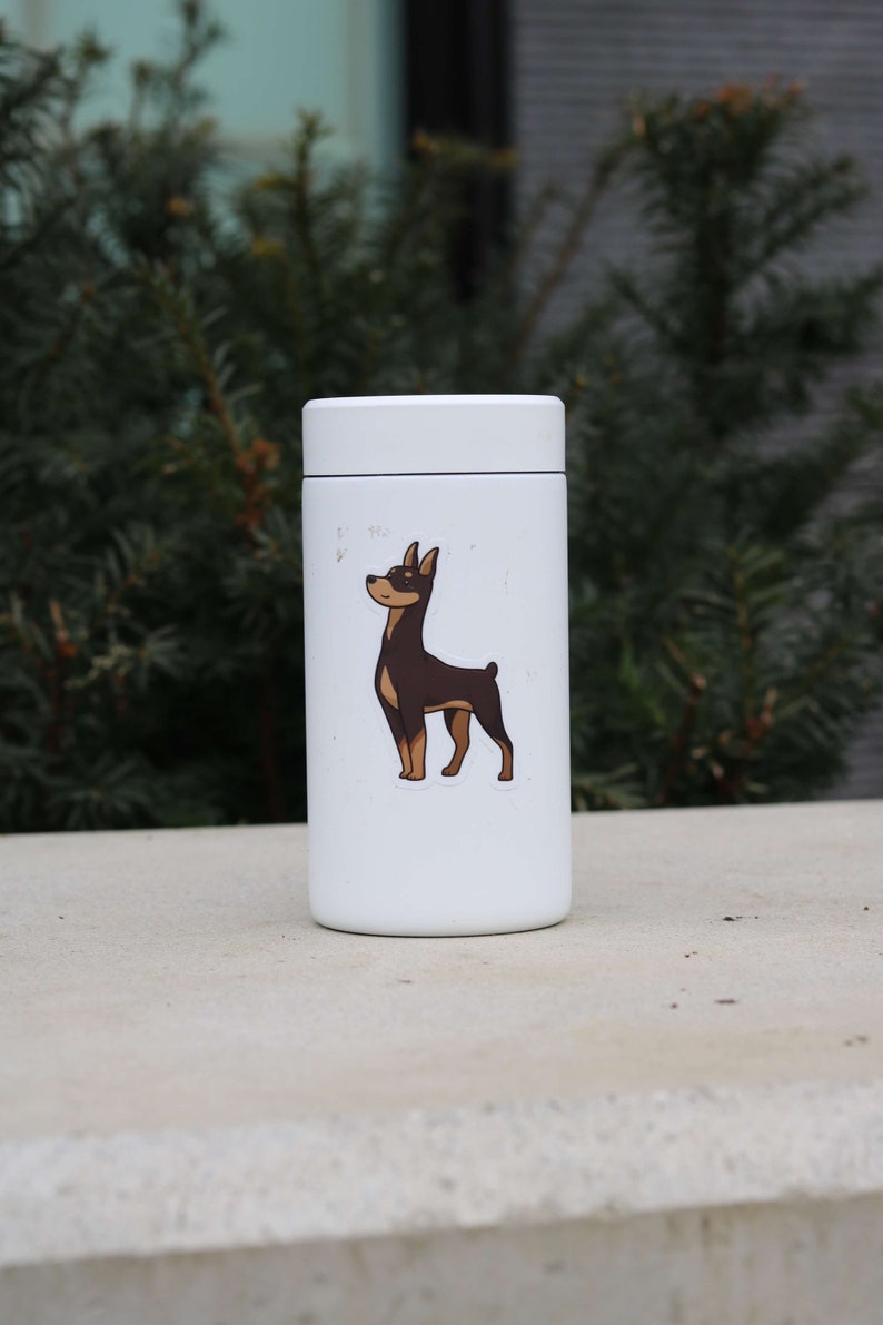 May include: White insulated water bottle with a brown and black dog sticker. The dog is looking up and has its tongue out.