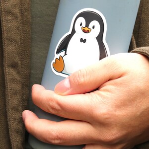 Puede incluir: Una botella de agua azul claro con una pegatina de ping&uuml;ino. El ping&uuml;ino es negro y blanco con patas naranjas. La botella la sostiene una persona que lleva una camisa verde y una chaqueta marr&oacute;n.
