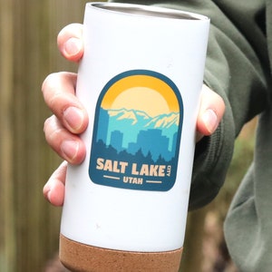 May include: White travel mug with a cork bottom and a sticker that says "Salt Lake City, Utah" with a graphic of a mountain range and a sun.