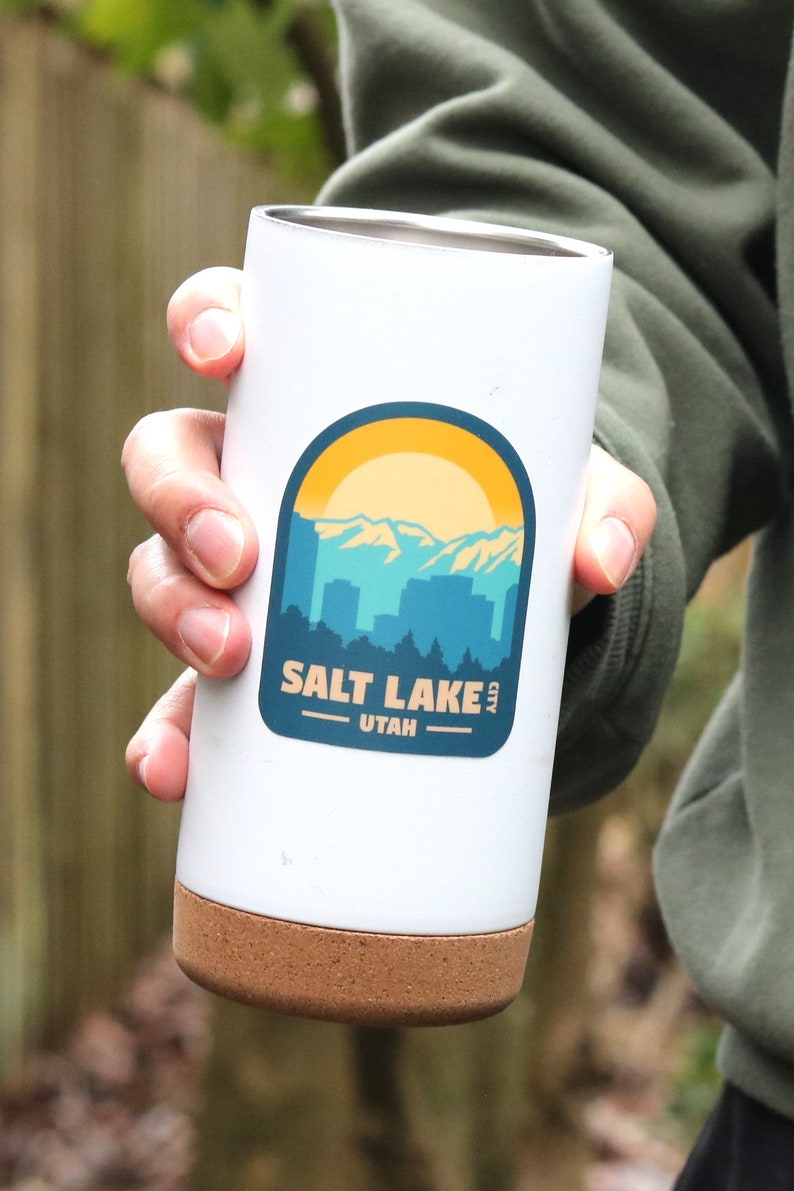 May include: White travel mug with a cork bottom and a sticker that says "Salt Lake City, Utah" with a graphic of a mountain range and a sun.