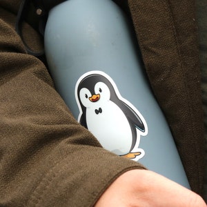 Puede incluir: Una botella de agua azul claro con tapa y asa negras. Una pegatina de un ping&uuml;ino de dibujos animados est&aacute; adherida a la botella. El ping&uuml;ino es blanco y negro con un pico naranja y una peque&ntilde;a pajarita negra. La botella la sostiene una persona con una chaqueta marr&oacute;n.