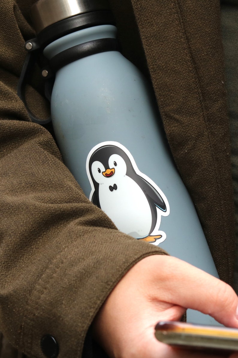 Puede incluir: Una botella de agua azul claro con tapa y asa negras. Una pegatina de un ping&uuml;ino de dibujos animados est&aacute; adherida a la botella. El ping&uuml;ino es blanco y negro con un pico naranja y una peque&ntilde;a pajarita negra. La botella la sostiene una persona con una chaqueta marr&oacute;n.