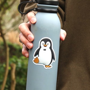 Puede incluir: Una botella de agua azul claro con un asa negra y una tapa plateada. Una pegatina de un ping&uuml;ino de dibujos animados con cuerpo negro, vientre blanco y patas naranjas est&aacute; en la botella.