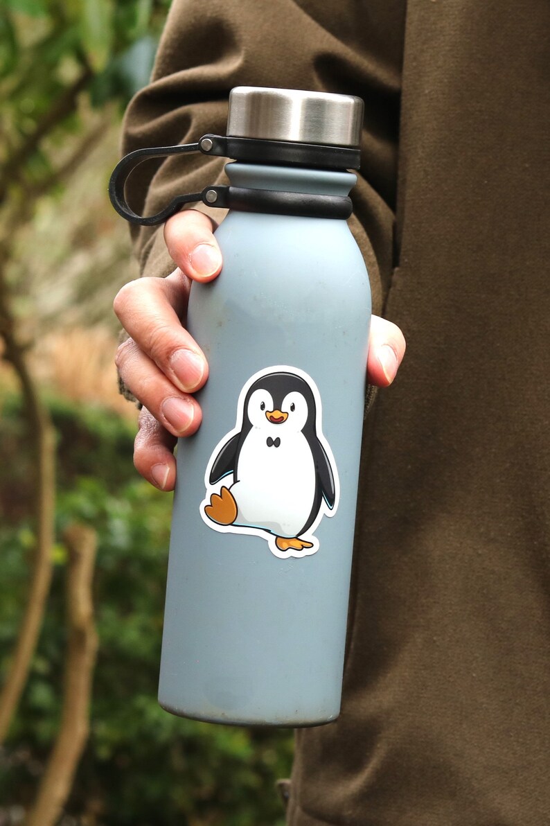 Puede incluir: Una botella de agua azul claro con un asa negra y una tapa plateada. Una pegatina de un ping&uuml;ino de dibujos animados con cuerpo negro, vientre blanco y patas naranjas est&aacute; en la botella.