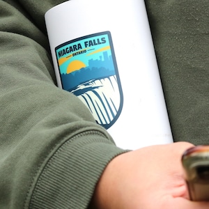 May include: White travel mug with a blue and green sticker that says "NIAGARA FALLS - ONTARIO" with a graphic of the falls and a yellow sun.
