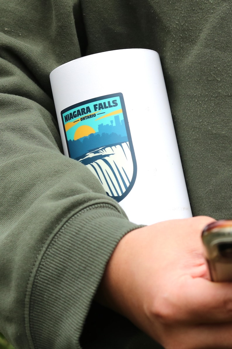 May include: White travel mug with a blue and green sticker that says "NIAGARA FALLS - ONTARIO" with a graphic of the falls and a yellow sun.
