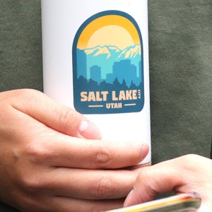 May include: White travel mug with a blue and yellow sticker featuring a cityscape and the text "SALT LAKE CITY UTAH".