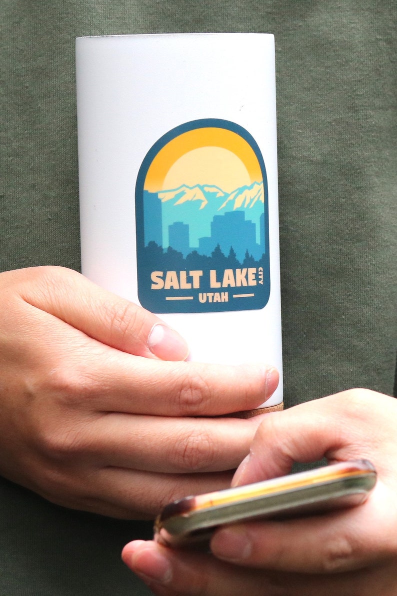 May include: White travel mug with a blue and yellow sticker featuring a cityscape and the text "SALT LAKE CITY UTAH".