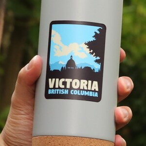 Puede incluir: Una taza de viaje gris con fondo de corcho. La taza tiene una pegatina con un cielo azul, nubes blancas, una silueta de un edificio y el texto "VICTORIA BRITISH COLUMBIA".