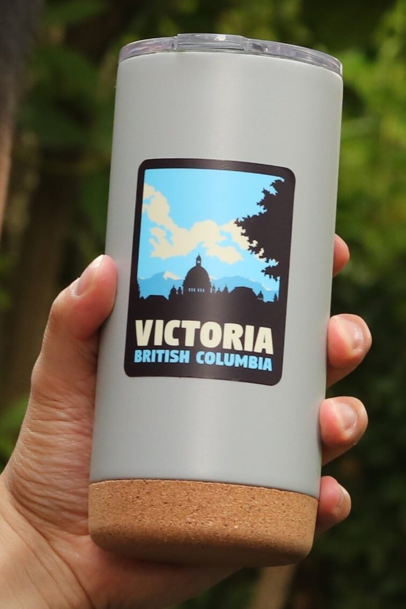 Puede incluir: Una taza de viaje gris con fondo de corcho. La taza tiene una pegatina con un cielo azul, nubes blancas, una silueta de un edificio y el texto "VICTORIA BRITISH COLUMBIA".