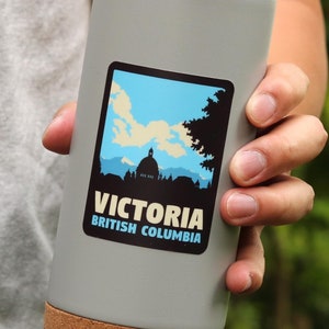 Puede incluir: Una taza de viaje gris con fondo de corcho y una pegatina que dice "Victoria, Columbia Brit&aacute;nica" con una silueta de un edificio y &aacute;rboles.