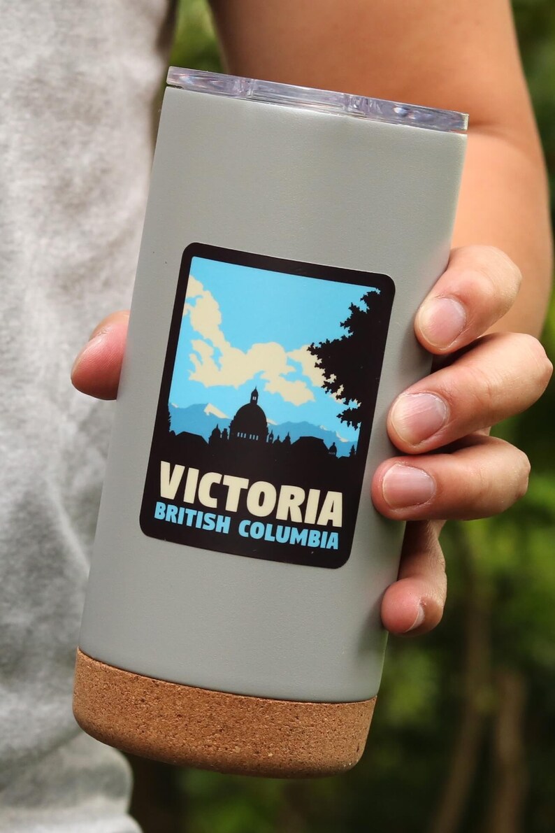 Puede incluir: Una taza de viaje gris con fondo de corcho y una pegatina que dice "Victoria, Columbia Brit&aacute;nica" con una silueta de un edificio y &aacute;rboles.