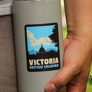 Puede incluir: Una taza de viaje gris con fondo de corcho. La taza tiene una pegatina con una silueta de un horizonte de ciudad y el texto "VICTORIA BRITISH COLUMBIA".