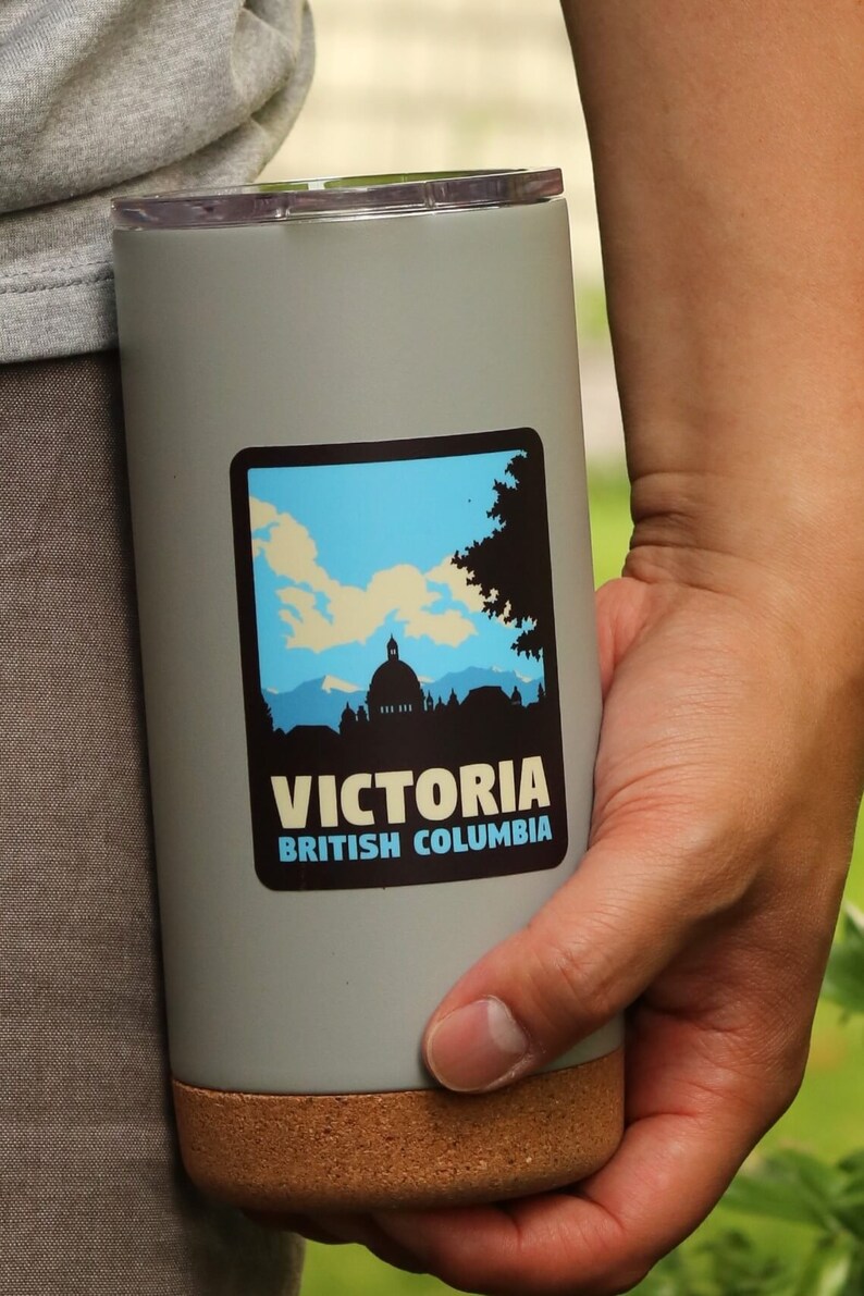 Puede incluir: Una taza de viaje gris con fondo de corcho. La taza tiene una pegatina con una silueta de un horizonte de ciudad y el texto "VICTORIA BRITISH COLUMBIA".