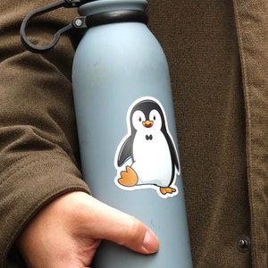 Puede incluir: Una botella de agua aislada de color azul claro con tapa plateada y asa negra. Una pegatina de ping&uuml;ino de dibujos animados est&aacute; adherida a la botella. El ping&uuml;ino es negro, blanco y naranja.