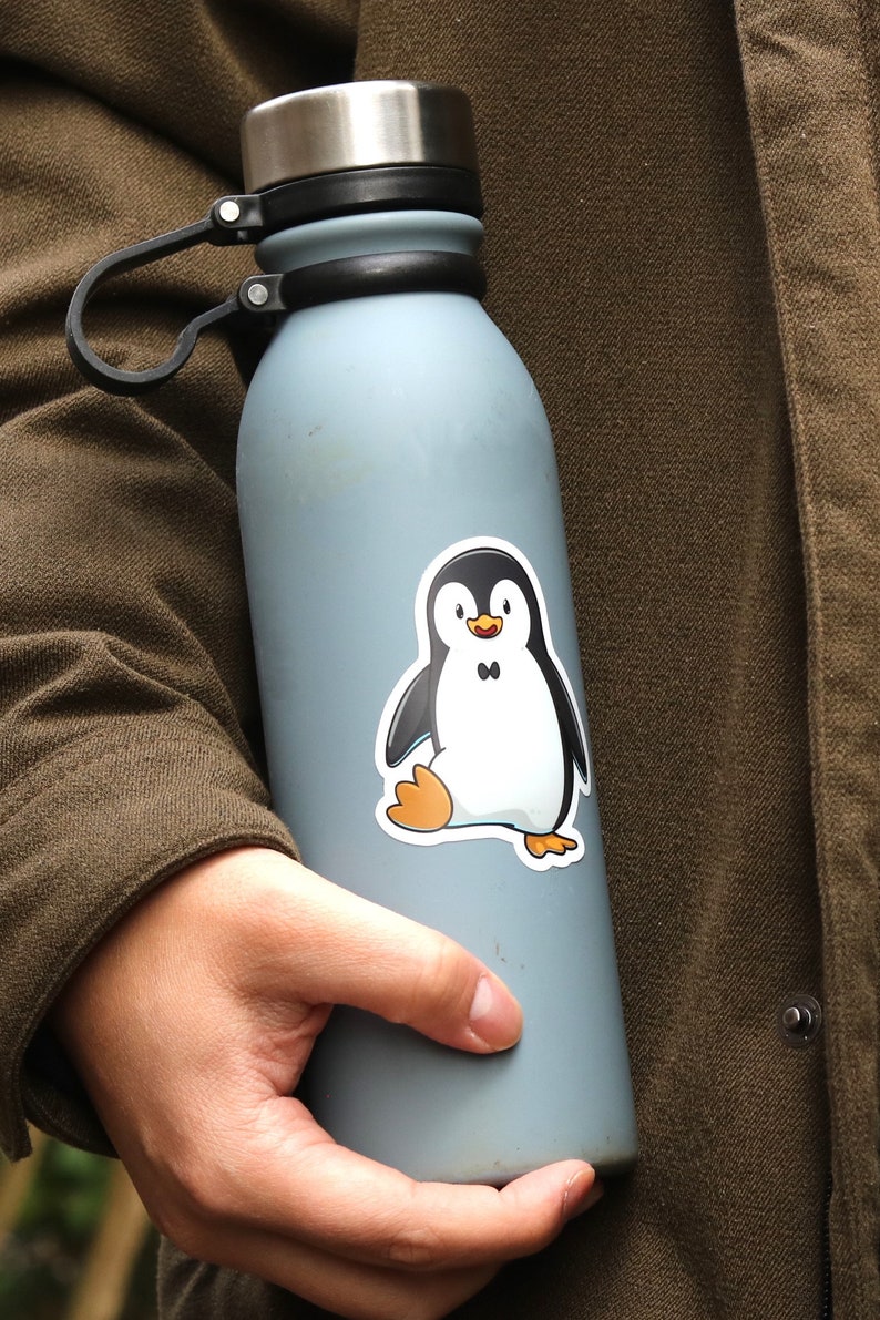 Puede incluir: Una botella de agua aislada de color azul claro con tapa plateada y asa negra. Una pegatina de ping&uuml;ino de dibujos animados est&aacute; adherida a la botella. El ping&uuml;ino es negro, blanco y naranja.