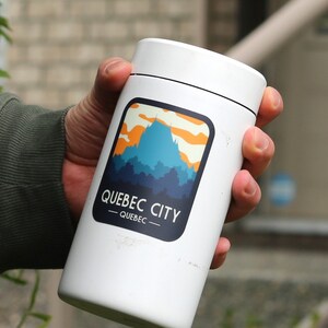 May include: A white cylindrical tumbler featuring a graphic illustration of a blue mountain silhouette against an orange and yellow sky. The text QUEBEC CITY and QUEBEC is printed in white within a dark blue rounded rectangular border on the front.