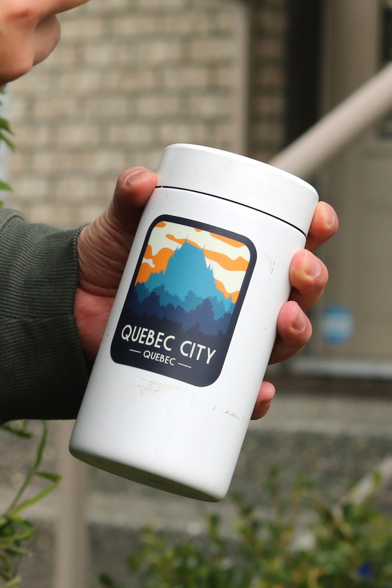 May include: A white cylindrical tumbler featuring a graphic illustration of a blue mountain silhouette against an orange and yellow sky. The text QUEBEC CITY and QUEBEC is printed in white within a dark blue rounded rectangular border on the front.