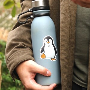 Puede incluir: Una botella de agua azul claro con un asa negra y una tapa plateada. Una pegatina de un ping&uuml;ino de dibujos animados con una pajarita negra est&aacute; en la botella. El ping&uuml;ino tiene un cuerpo blanco, alas negras y pies naranjas.