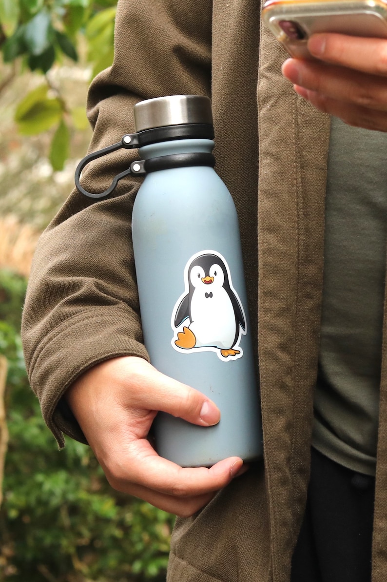 Puede incluir: Una botella de agua azul claro con un asa negra y una tapa plateada. Una pegatina de un ping&uuml;ino de dibujos animados con una pajarita negra est&aacute; en la botella. El ping&uuml;ino tiene un cuerpo blanco, alas negras y pies naranjas.