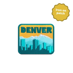 Peut inclure: Patch thermocollant représentant une ville avec le mot "DENVER" en texte vert sur fond jaune. La ville est soulignée en turquoise et présente des montagnes blanches en arrière-plan.
