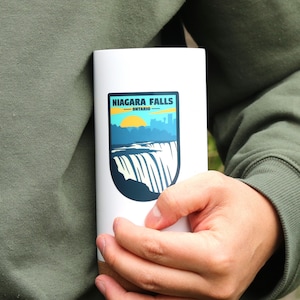May include: A white travel mug with a brown wood bottom. The mug has a sticker on the front that says "NIAGARA FALLS - ONTARIO" with a graphic of the falls and a sunset.