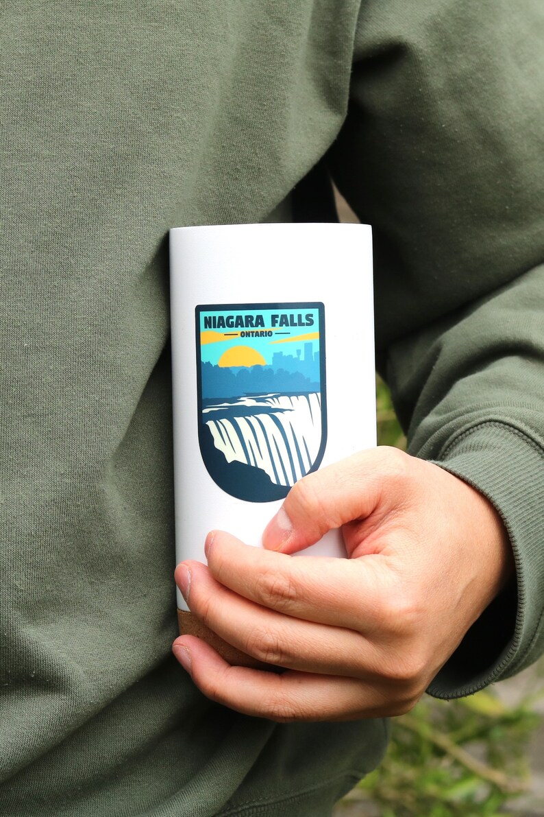 May include: A white travel mug with a brown wood bottom. The mug has a sticker on the front that says "NIAGARA FALLS - ONTARIO" with a graphic of the falls and a sunset.