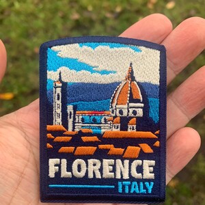 K&ouml;nnte beinhalten: Gestickter Aufn&auml;her mit der Kathedrale von Florenz in Italien. Der Aufn&auml;her hat einen blauen Hintergrund mit wei&szlig;en, orangefarbenen und blauen architektonischen Details. Der Text "FLORENCE ITALY" ist wei&szlig; gestickt.