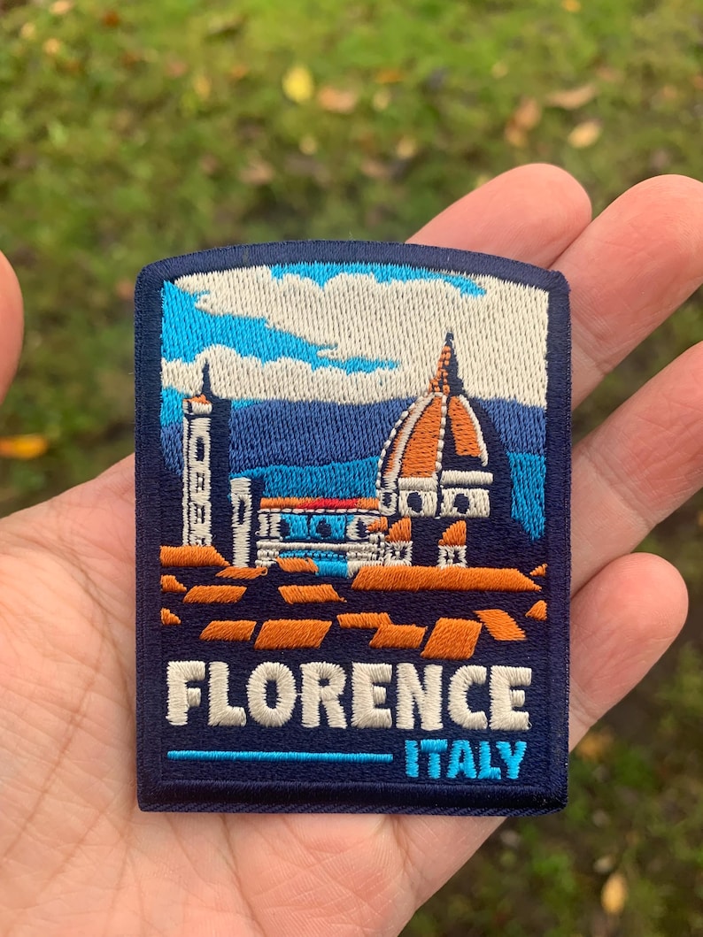 K&ouml;nnte beinhalten: Gestickter Aufn&auml;her mit der Kathedrale von Florenz in Italien. Der Aufn&auml;her hat einen blauen Hintergrund mit wei&szlig;en, orangefarbenen und blauen architektonischen Details. Der Text "FLORENCE ITALY" ist wei&szlig; gestickt.