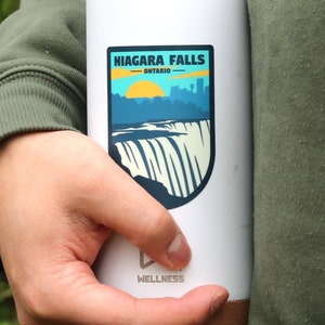 May include: White travel mug with a brown bottom. The mug has a sticker on the front that says "Niagara Falls - Ontario" with a graphic of the falls and a sunset. The bottom of the mug says "Wellness".