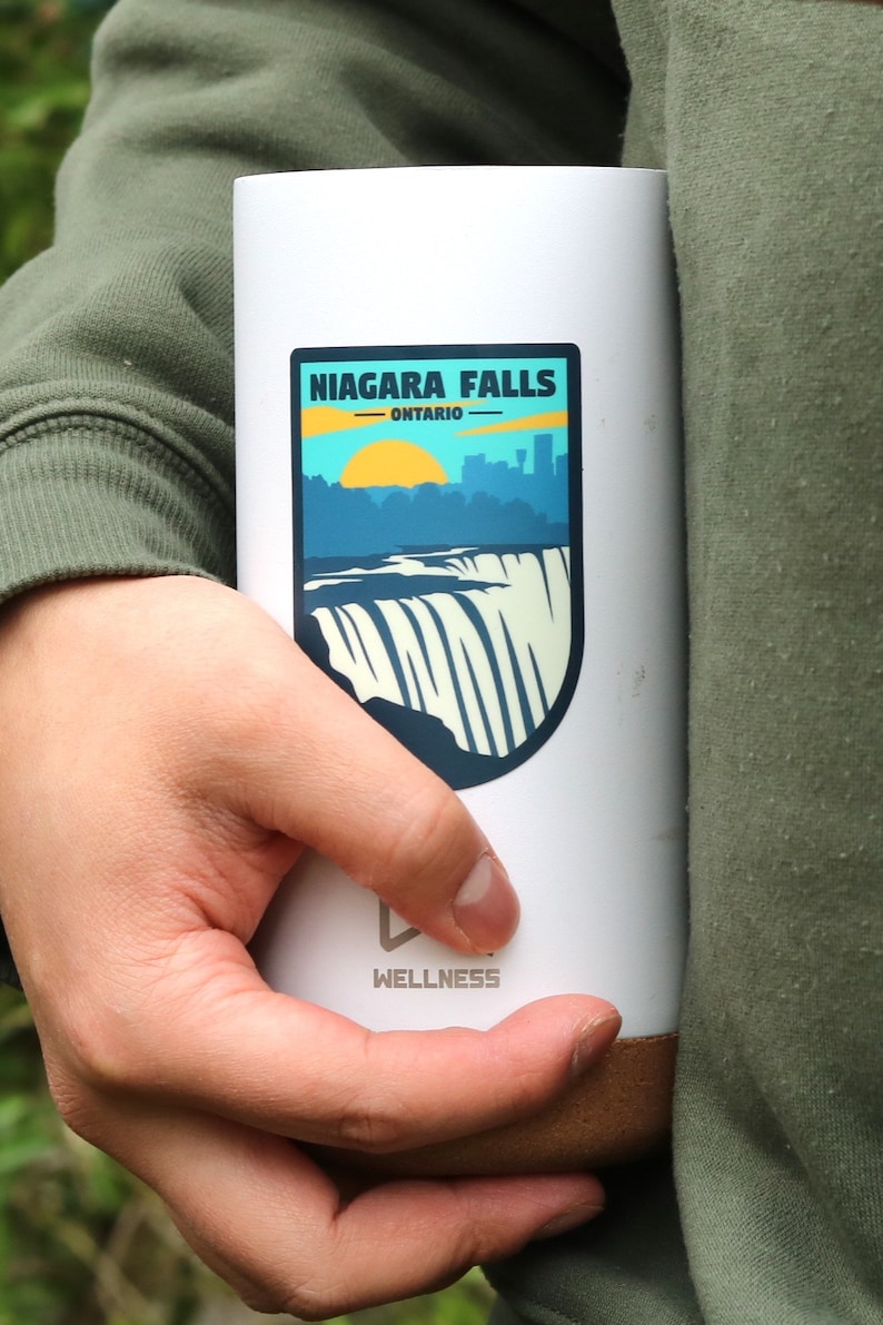 May include: White travel mug with a brown bottom. The mug has a sticker on the front that says "Niagara Falls - Ontario" with a graphic of the falls and a sunset. The bottom of the mug says "Wellness".
