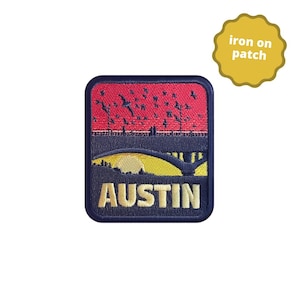 May include: An iron-on patch featuring a stylized landscape with the word "AUSTIN" in gold lettering. The design includes a red sky, dark blue hills, and a yellow sun. The patch is rectangular with rounded corners.