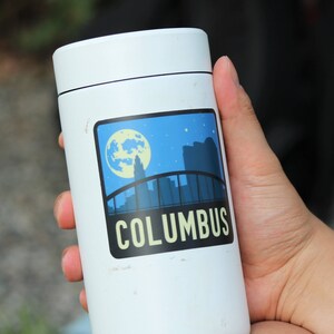 May include: White insulated travel mug with a sticker featuring a skyline and the word "COLUMBUS" in yellow letters.