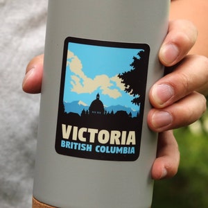 Puede incluir: Una taza de viaje de acero inoxidable gris con fondo de corcho. La taza tiene una pegatina en la parte delantera que dice "Victoria, Columbia Brit&aacute;nica" y presenta una silueta de un horizonte de ciudad con un edificio en forma de c&uacute;pula.