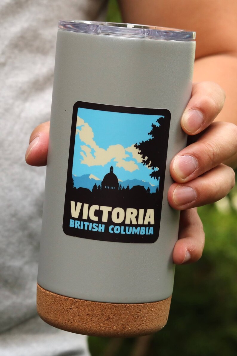 Puede incluir: Una taza de viaje de acero inoxidable gris con fondo de corcho. La taza tiene una pegatina en la parte delantera que dice "Victoria, Columbia Brit&aacute;nica" y presenta una silueta de un horizonte de ciudad con un edificio en forma de c&uacute;pula.